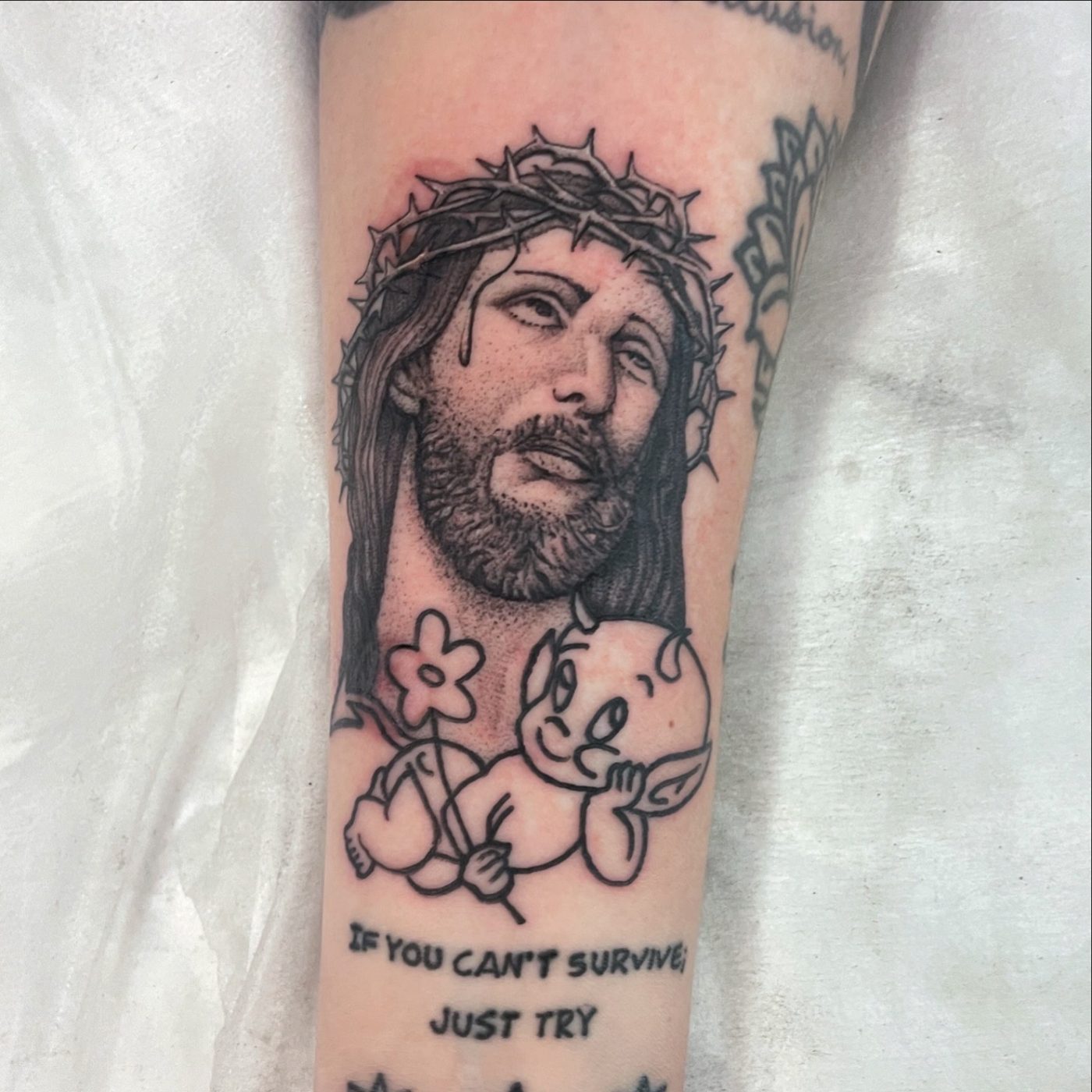 Black and grey arm tattoo of Jesus in a crown of thorns above a small outlined cherub holding a flower