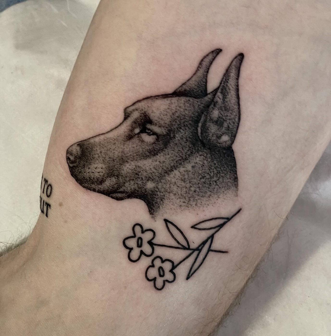 Dotwork arm tattoo portrait of a dog in profile above two simple outlined flowers