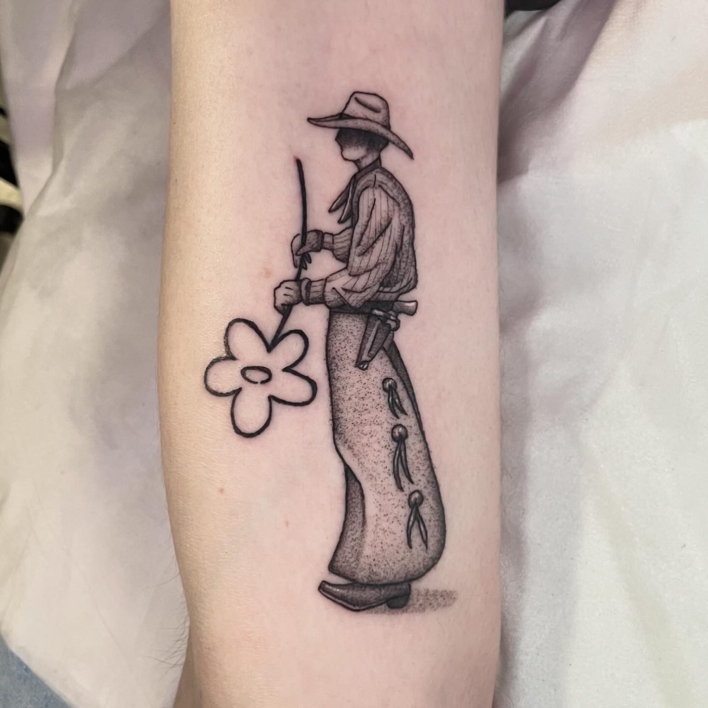 Fine-line arm tattoo of a cowboy in chaps and a wide hat cutting a single outlined flower
