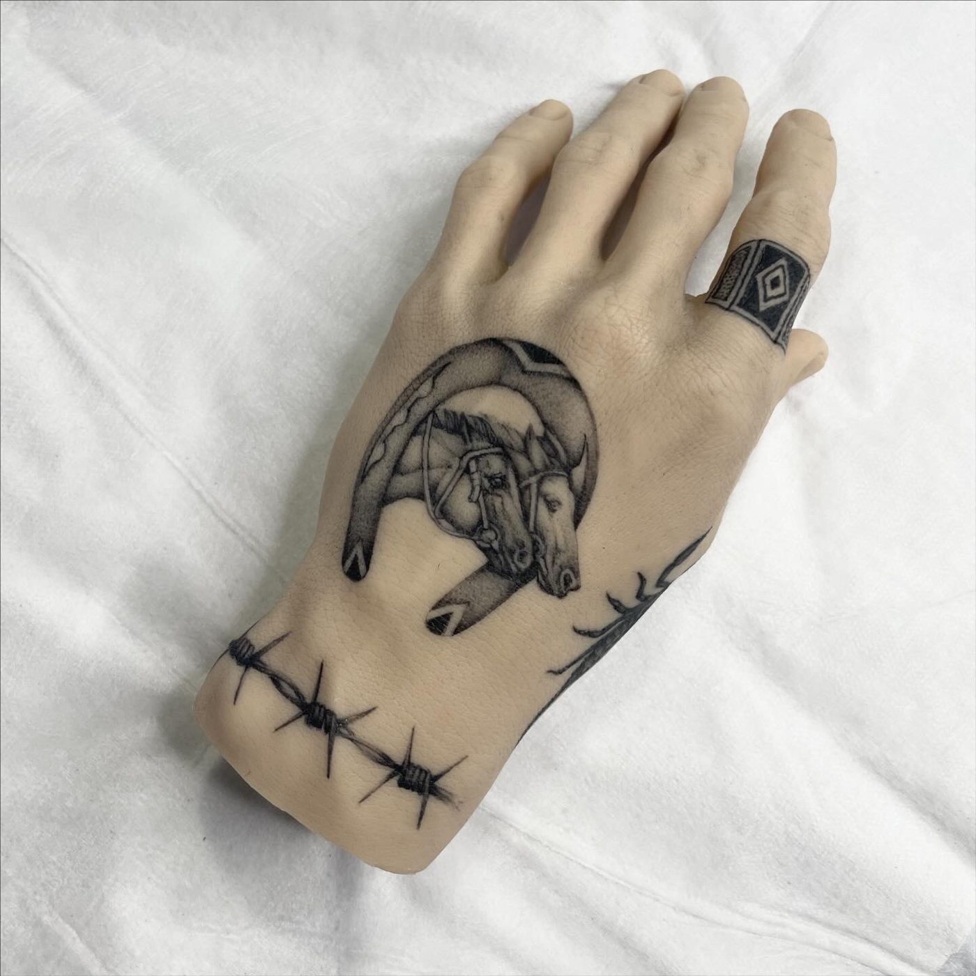 Hand tattoo of two horse heads inside a horseshoe, with a barbed wire band across the wrist